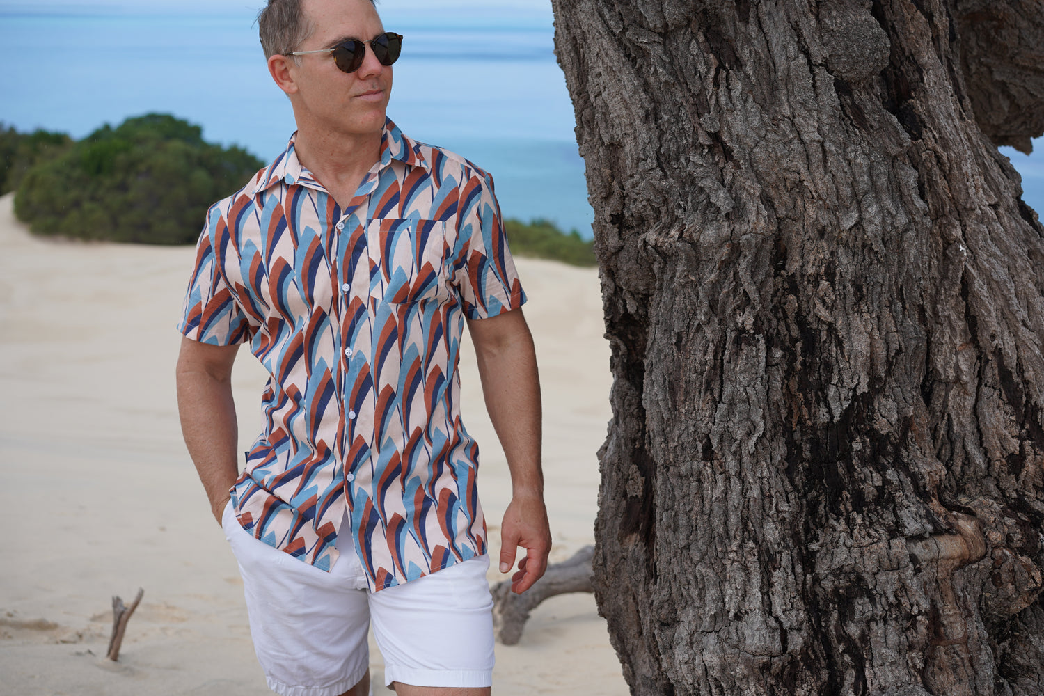 Image of man standing next to tree on sand dune with ocean in background wearing BroadReach resort wear Helmsman short sleeved button-up shirt in Liberty Fabrics Refracted Light print.
