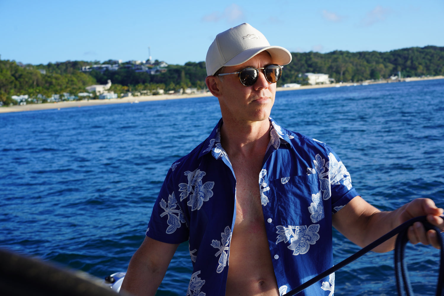 Image of man standing yacht with island in background wearing BroadReach resort wear Helmsman short sleeved button-up shirt in Liberty Fabrics Petal Patterns print and BroadReach Sundowner cap in sand color.