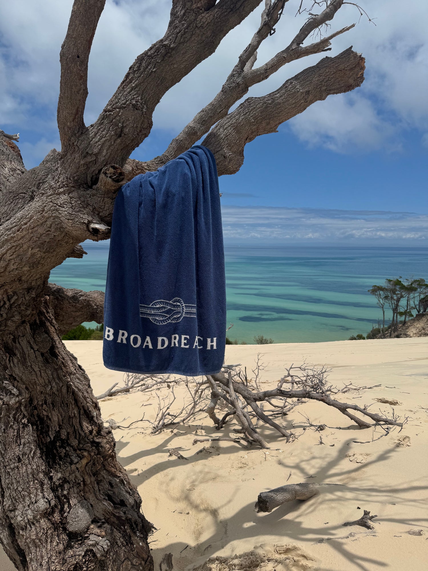 Image of Poolside towel by BroadReach resort wear hanging on tree trunk with sand dune and ocean in background.